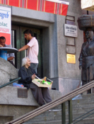 An old woman begs by offering vegetables for sale outside the market, supervised by a sturdy professional idealized in bronze
